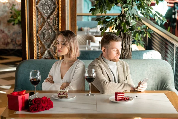 年轻无聊或生气的女人坐在餐厅的餐桌旁 男朋友在旁边的智能手机里打滚 — 图库照片