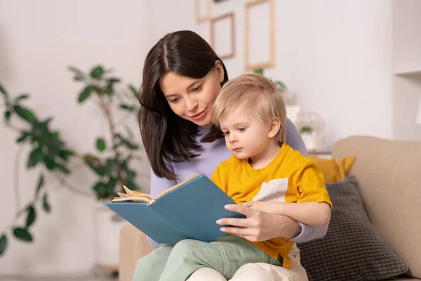 説明美しいです若いです母座っていますソファの上にリビングルームでとブロンドの髪の息子へのおとぎ話を読みます — ストック写真