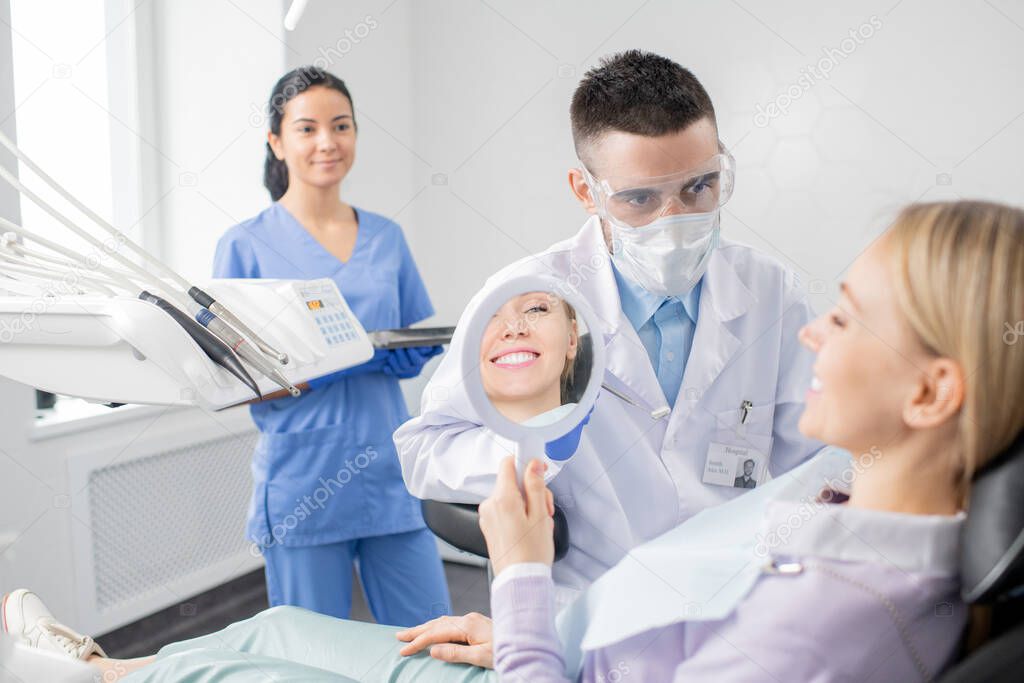 Happy young female patient with toothy smile looking in mirror while sitting in armchair in dentistry office with her dentist in front