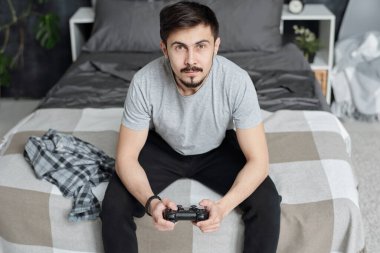 Evde karantina sırasında oyun oynarken yatakta oturup joystick kullanan meraklı genç adam.