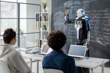 İki genç öğrenci, modern teknolojinin dersinde masanın başında oturmuş, yeteneklerini karatahta ile gösteren robotlara bakıyorlar.