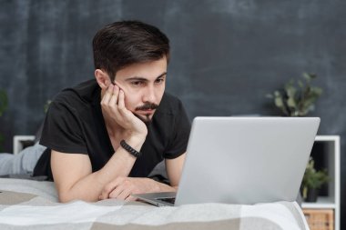 Ciddi yakışıklı genç adam yatakta uzanmış kendini izole bir şekilde internetten film izliyor.