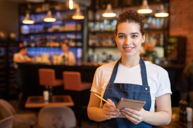 İş kıyafetleri içinde genç bir garson lüks bir restoranda kameranın önünde duruyor ve müşteri sırasına göre yazışıyor.