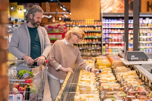 Happy mature female taking packed piece of cheese from display while walking along dairy products with her husband in supermarket
