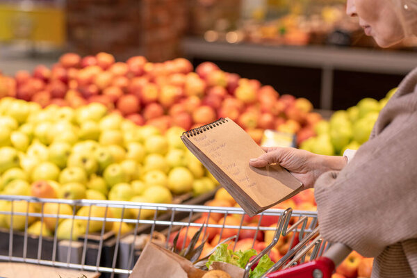 Mature female consumer with notepad over food products in cart looking through shopping list while moving along fruit display