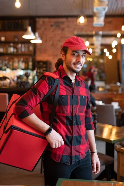 Young courier in red cap and flannel carrying big bag on shoulder while going to leave restaurant and deliver ordered food to clients