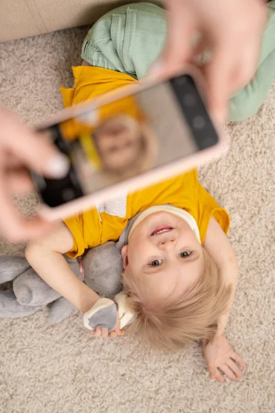 床に玩具を寝そべって幸せなかわいい息子の写真を撮っている間にスマートフォンを使用して認識できない親の上記のビュー — ストック写真
