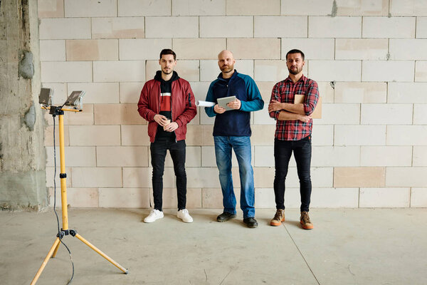 Three young male engineers or builders in workwear standing in line along wall of unfinished building in front of camera during work