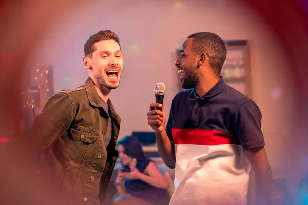 Two cheerful intercultural guys singing together in microphone while enjoying karaoke party on background of their girlfriends
