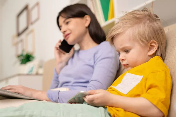 かわいいですブロンド男の子とともにスマートフォンオンライン漫画を見ている間彼の母親は電話で話していると隔離にリモートで作業 — ストック写真
