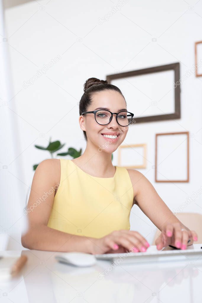 Portrait of smiling beautiful young businesswoman in eyeglasses sitting at desk in modern office and typing on computer