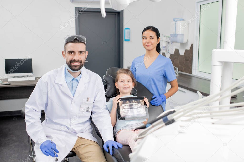 Happy young dentist in whitecoat, his little patient and assistant in uniform looking at you by workplace in contemporary dental clinics