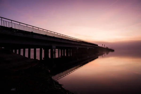 Largo puente diagonal y río al amanecer en la madrugada en una neblina de niebla, naturaleza y civilización —  Fotos de Stock