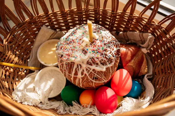 Easter straw basket decorated with a festive napkin with Easter cake and bright decorated eggs and a lit church candle. A festive treat and a peaceful mood.