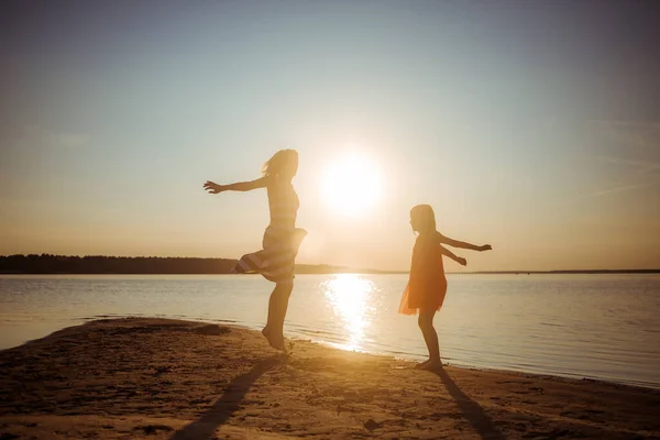 Happy mom and daughter in dresses are jumping and dancing on the beach during sunset. Good relations of two generations. Fun family pastime in nature. Health promotion through games and outdoor activities.