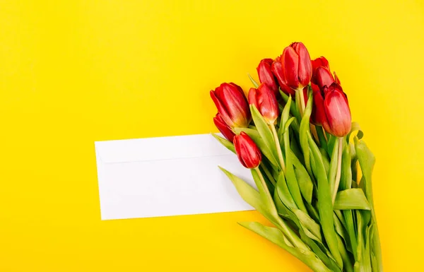 White gift envelope on a yellow background with a bouquet of red tulips and a free place for text. Sample certificate or invitation card. Close-up.