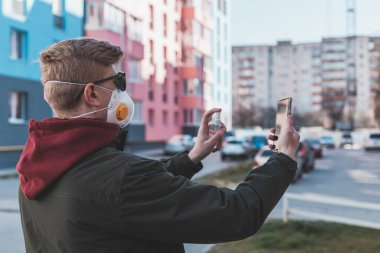 Gözlüklü ve koruyucu maskeli bir adam virüs salgınının karantinası sırasında akıllı telefonunu sokakta dezenfektanla temizliyor..