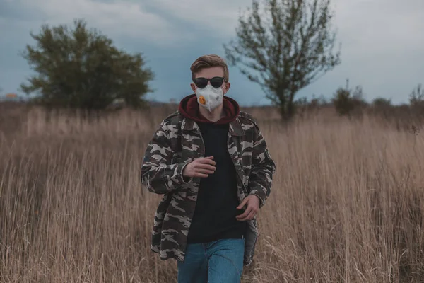 A man in a protective mask and camouflage jacket runs across the field in nature. Jogging during self-isolation during the coronavirus epidemic.
