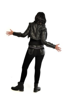 tam uzunlukta uzak kameradan bakan deri kıyafeti giymiş siyah saçlı kız portresi. Beyaz stüdyo arka plan üzerinde ayakta poz.