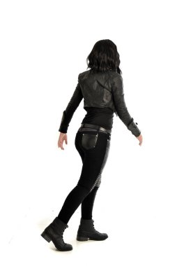 tam uzunlukta uzak kameradan bakan deri kıyafeti giymiş siyah saçlı kız portresi. Beyaz stüdyo arka plan üzerinde ayakta poz.