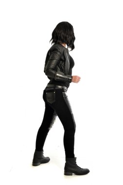 tam uzunlukta uzak kameradan bakan deri kıyafeti giymiş siyah saçlı kız portresi. Beyaz stüdyo arka plan üzerinde ayakta poz.