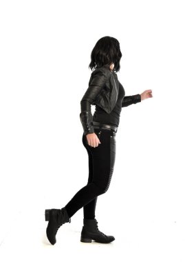 tam uzunlukta uzak kameradan bakan deri kıyafeti giymiş siyah saçlı kız portresi. Beyaz stüdyo arka plan üzerinde ayakta poz.