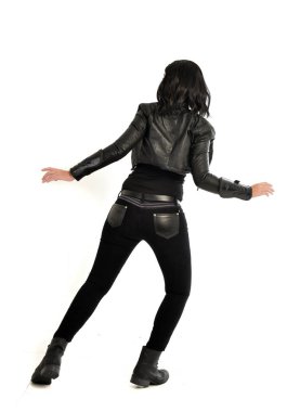 tam uzunlukta uzak kameradan bakan deri kıyafeti giymiş siyah saçlı kız portresi. Beyaz stüdyo arka plan üzerinde ayakta poz.
