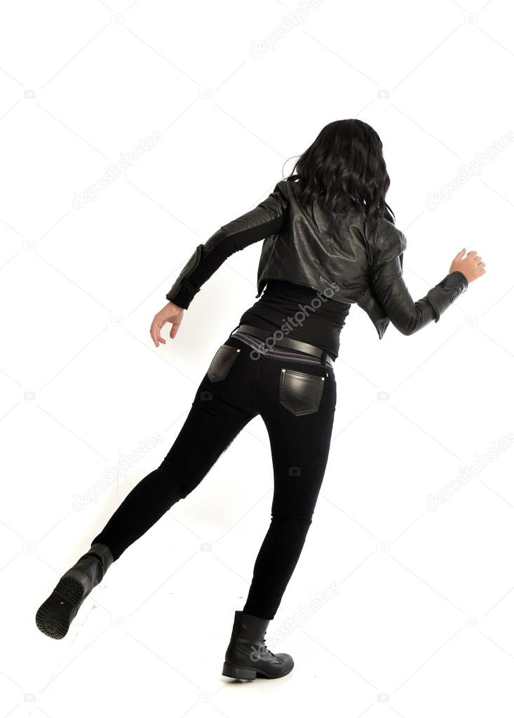 full length portrait of black haired girl wearing leather outfit, facing away from the camera. standing pose on a white studio background.