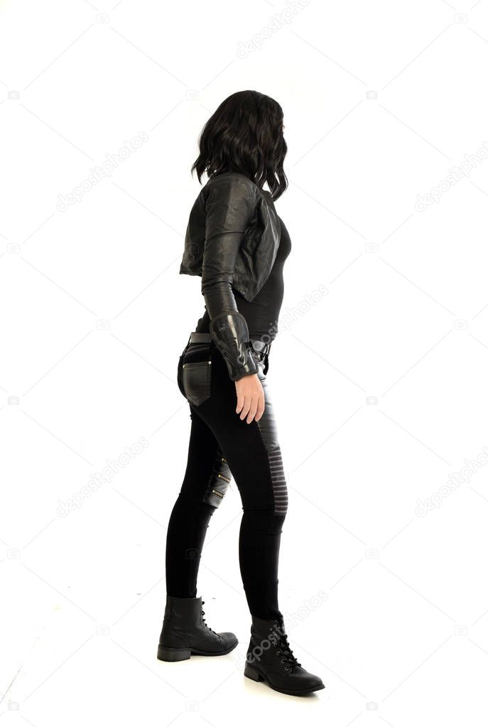 full length portrait of black haired girl wearing leather outfit, facing away from the camera. standing pose on a white studio background.