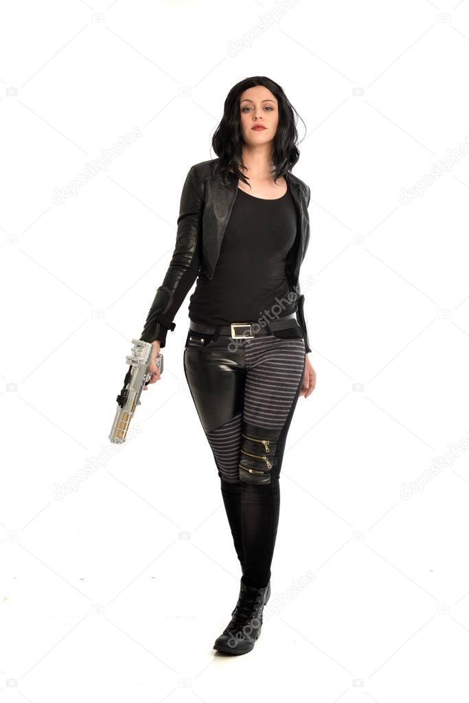 full length portrait of black haired girl wearing leather outfit. standing pose while holding a gun, isolated on a white studio background.