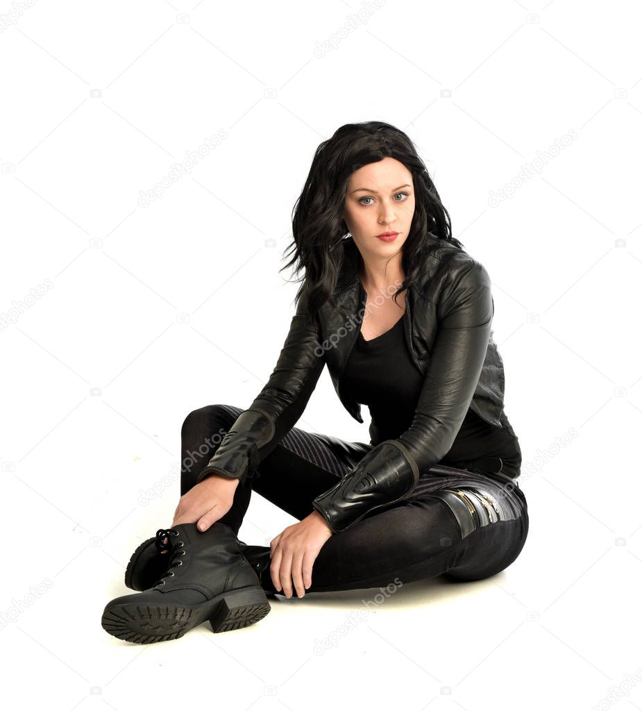 full length portrait of black haired girl wearing leather outfit.  seated on pose, isolated on a white studio background.