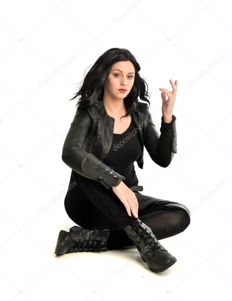 full length portrait of black haired girl wearing leather outfit.  seated on pose, isolated on a white studio background.