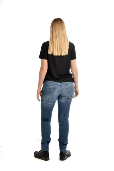 シンプルな黒シャツとジーンズを身に着けているブロンドの女の子の完全な長さの肖像画 白い背景で隔離のカメラから離れて直面して立ちポーズ — ストック写真