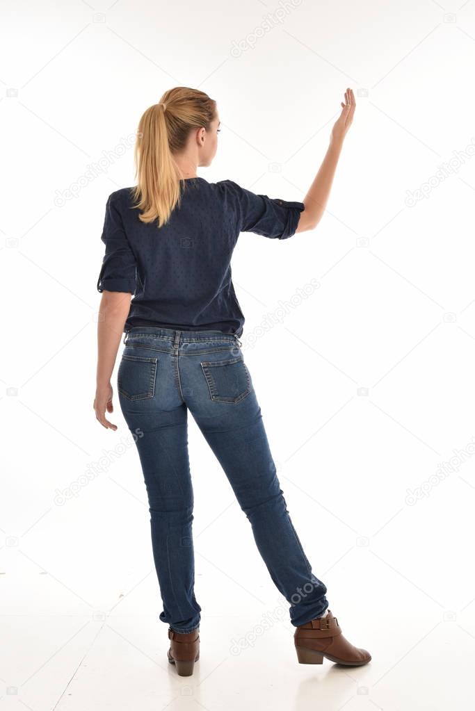 full length portrait of blonde girl wearing simple blue shirt and jeans, standing pose facing away from the camera. isolated on white background.