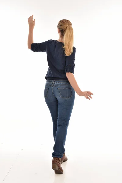カメラから離れて直面しているポーズに立っているシンプルなブルーのシャツとジーンズを身に着けている金髪の女の子の完全な長さの肖像画 白い背景に分離 — ストック写真