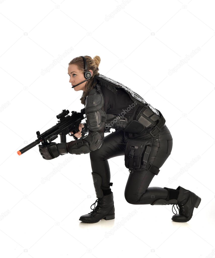 full length portrait of female  soldier wearing black  tactical armour, seated pose holding a gun, isolated on white studio background.