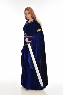 tam uzunlukta bir mavi kadife Ortaçağ elbisesi ve kürk Pelerin giyen kadın uzun saçlı portresi. ayakta poz beyaz arka plan üzerinde izole uzun bir kılıç holding.