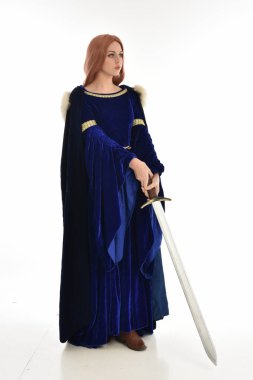 tam uzunlukta bir mavi kadife Ortaçağ elbisesi ve kürk Pelerin giyen kadın uzun saçlı portresi. ayakta poz beyaz arka plan üzerinde izole uzun bir kılıç holding.