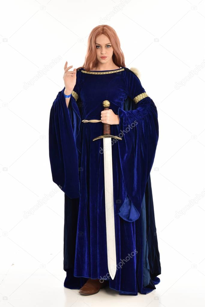 full length portrait of woman with long hair, wearing a blue velvet medieval gown and fur cloak. standing pose holding a long sword, isolated on white background.