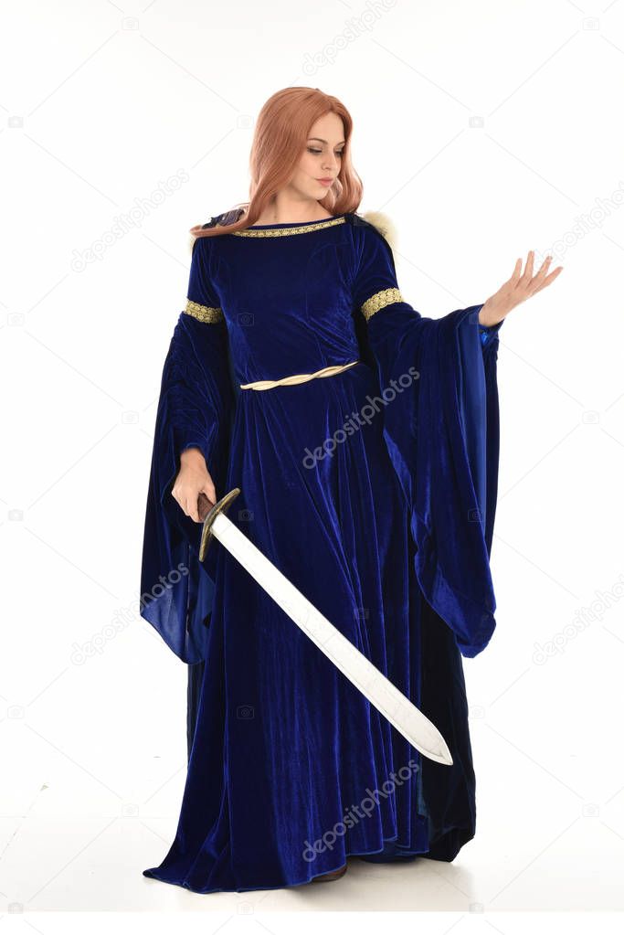 full length portrait of woman with long hair, wearing a blue velvet medieval gown and fur cloak. standing pose holding a long sword, isolated on white background.
