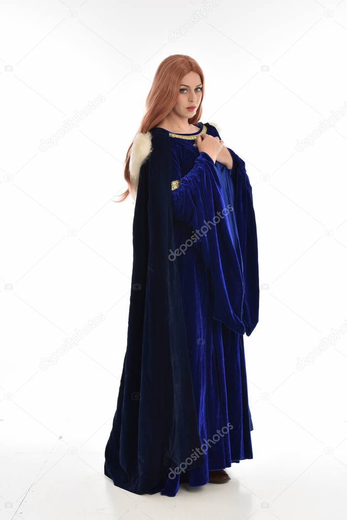 full length portrait of woman with long hair, wearing a blue velvet medieval gown and fur cloak. standing pose in side profile, isolated on white background.
