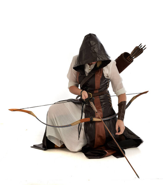 full length portrait of lad wearing brown leather fantasy costume, kneeling pose holding a bow and arrow.  isolated on a white studio background.
