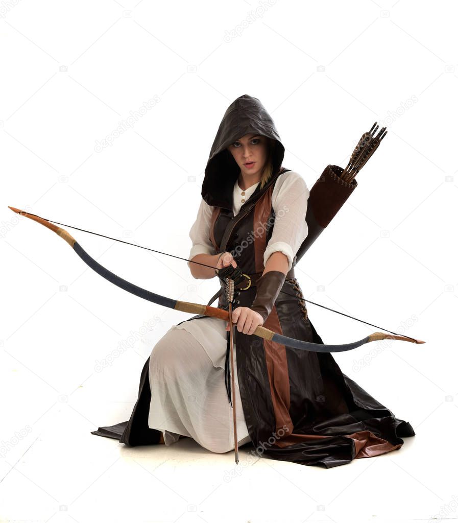 full length portrait of lad wearing brown leather fantasy costume, kneeling pose holding a bow and arrow.  isolated on a white studio background.