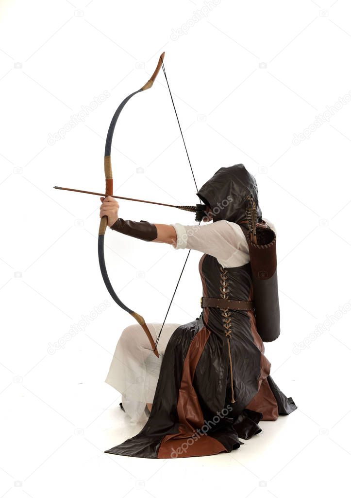full length portrait of lad wearing brown leather fantasy costume, kneeling pose holding a bow and arrow.  isolated on a white studio background.
