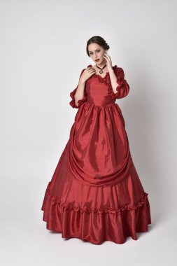 Kırmızı ipek Victoria elbisesi giyen esmer bir kızın tam boy portresi. Beyaz bir stüdyonun arka planında poz vermek.