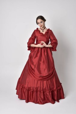 Kırmızı ipek Victoria elbisesi giyen esmer bir kızın tam boy portresi. Beyaz bir stüdyonun arka planında poz vermek.