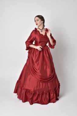 full length portrait of a brunette girl wearing a red silk victorian gown. Standing pose on a white studio background. clipart