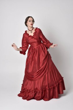 Kırmızı ipek Victoria elbisesi giyen esmer bir kızın tam boy portresi. Beyaz bir stüdyonun arka planında poz vermek.