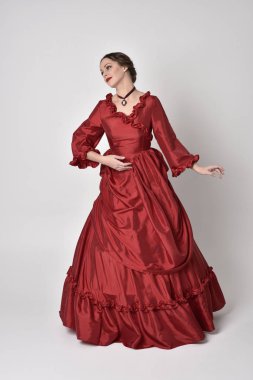 Kırmızı ipek Victoria elbisesi giyen esmer bir kızın tam boy portresi. Beyaz bir stüdyonun arka planında poz vermek.