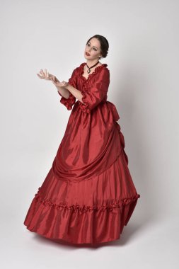 Kırmızı ipek Victoria elbisesi giyen esmer bir kızın tam boy portresi. Beyaz bir stüdyonun arka planında poz vermek.